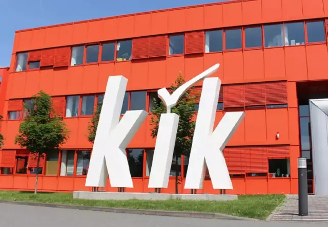 Modernes Gebäude mit leuchtend roter Fassade mit dem großen weißen „kik“-Logo und Eingang, umgeben von Grün an einem sonnigen Tag