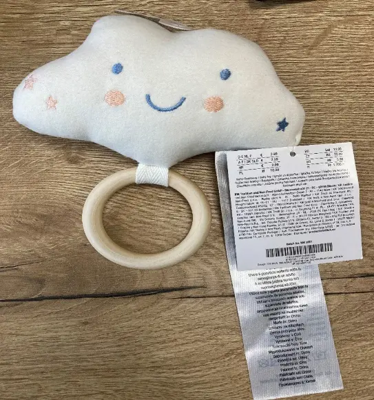 Ein weiches, plüschiges Babyspielzeug in Wolkenform mit lächelndem Gesicht, Holzring und angenähtem Pflegeetikett auf einer Holzoberfläche. Ein weiches, plüschiges Babyspielzeug in Wolkenform mit lächelndem Gesicht, Holzring und angenähtem Pflegeetikett auf einer Holzoberfläche.