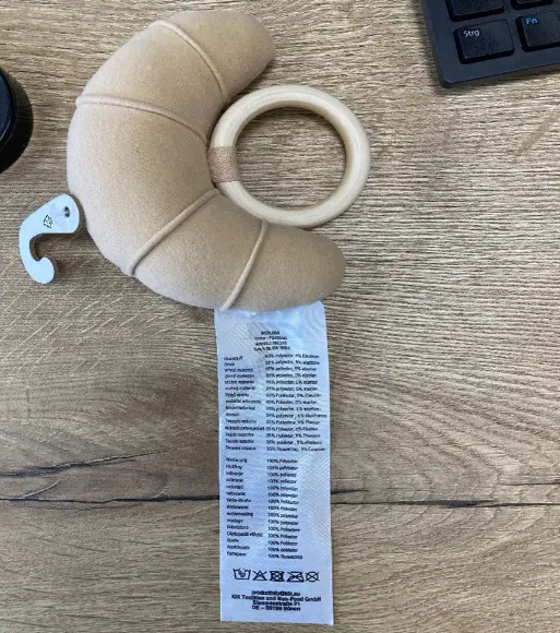 Ein weiches, croissantförmiges Plüschtier mit Holzring und Pflegeetikett, auf einer Holzoberfläche neben einer Tastatur liegend. Ein weiches, croissantförmiges Plüschtier mit Holzring und Pflegeetikett, auf einer Holzoberfläche neben einer Tastatur liegend.