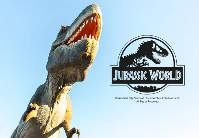 Jurassic World - Kinderkleidung Ein großer, brüllender T-Rex erhebt sich vor dem wolkenlosen Himmel, und das Logo von Jurassic World ist gut sichtbar.