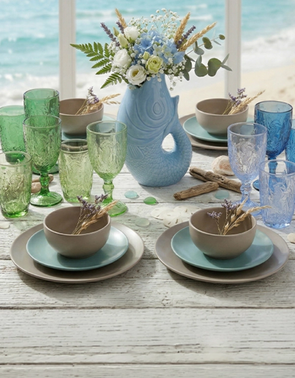 Maritim gedeckter Tisch mit farbigen Gläsern, Geschirr und dekorativer Vase mit Blumen vor Meereskulisse – stilvolle Tischdekoration für sommerliche Wohlfühlmomente
