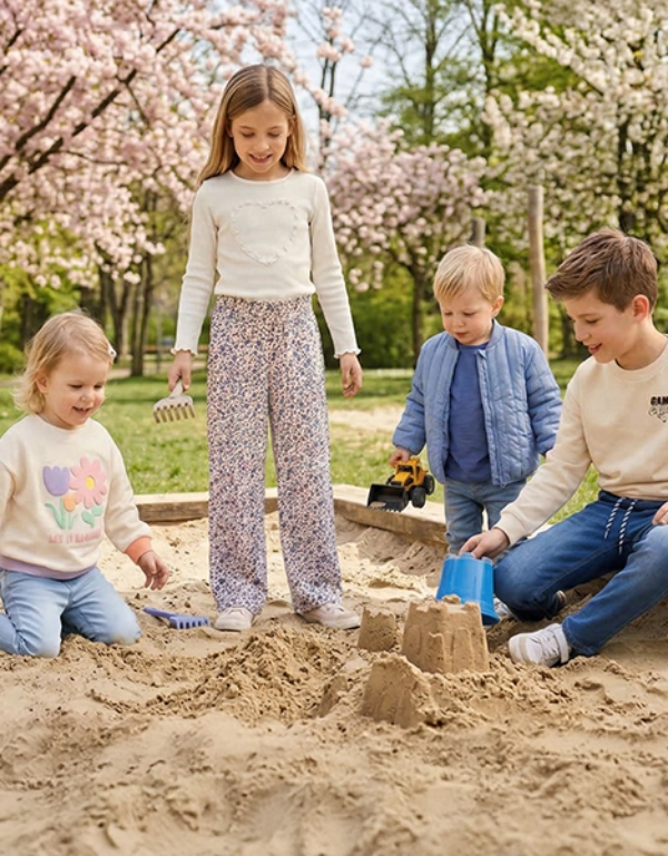 Vier Kinder spielen und bauen Sandburgen in einem Sandkasten unter blühenden Bäumen in einem Park.