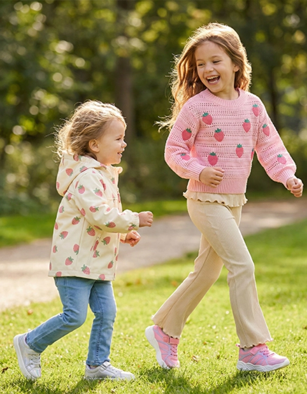 Zwei Kinder laufen fröhlich durch einen sonnendurchfluteten Park, umgeben von grünem Gras und Bäumen, und tragen bunte Pullover und Freizeithosen.