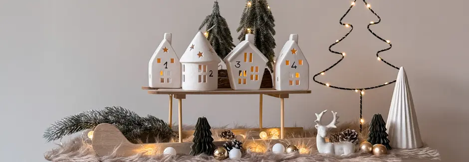 Eine festliche Dekoration aus vier Keramikhäusern mit ausgeschnittenen Fenstern, umgeben von kleinen Bäumen, Ornamenten und Lichterketten.