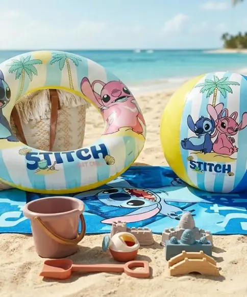 Strandtuch, Schwimmring, Ball und Sandspielzeug mit Lilo‑&‑Stitch‑Motiv liegen am Sandstrand neben türkisfarbenem Meer.
