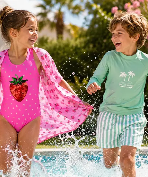 Zwei Kinder laufen am Pool, eines trägt einen pinken Badeanzug mit Erdbeermotiv und ein pinkes Überwurfkleid, das andere ein grünes Langarmshirt mit Palmenmotiv und grün-weiß gestreifte Badeshorts