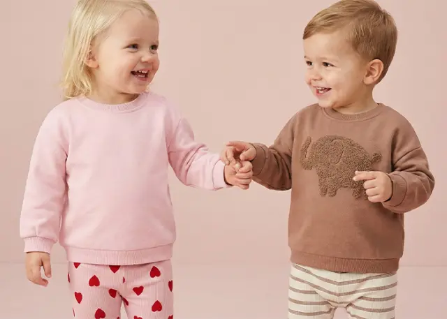 Zwei kleine Kinder halten sich an den Händen, eines trägt einen rosa Pullover und gestreifte Hosen mit Herzmuster, das andere einen braunen Pullover mit Elefantenmotiv. Zwei kleine Kinder halten sich an den Händen, eines trägt einen rosa Pullover und gestreifte Hosen mit Herzmuster, das andere einen braunen Pullover mit Elefantenmotiv.