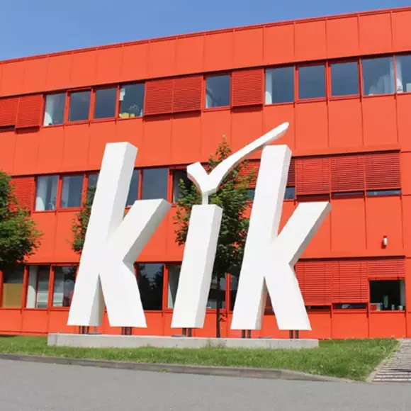 KiK Logo vor einem Gebäude Modernes Gebäude mit leuchtend roter Fassade mit dem großen weißen „kik“-Logo und Eingang, umgeben von Grün an einem sonnigen Tag