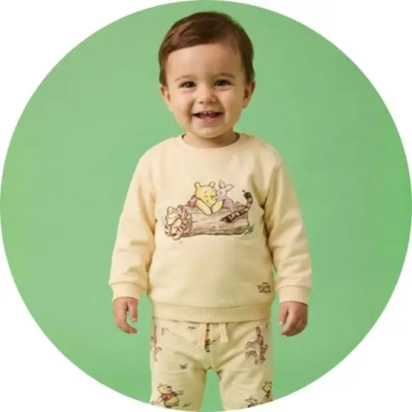 Ein Kleinkind steht vor grünem Hintergrund und trägt einen gelben Pullover mit einer verspielten Illustration von Comicfiguren.