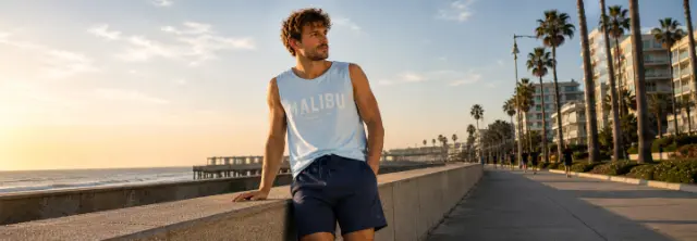 Herren Sommeroutfit mit hellblauem Tanktop und dunkelblauen Shorts an der Strandpromenade bei Sonnenuntergang