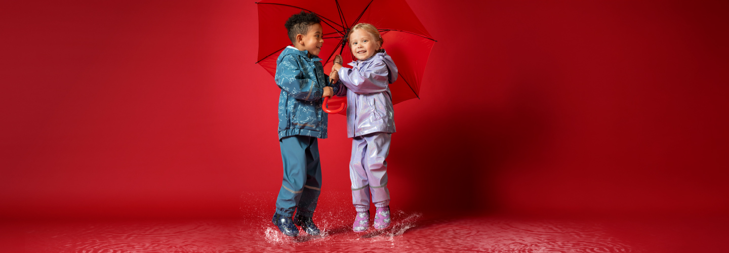 Zwei Kinder in bunten Regenmänteln planschen fröhlich in Pfützen, während sie sich einen leuchtend roten Regenschirm teilen. Zwei Kinder in bunten Regenmänteln planschen fröhlich in Pfützen, während sie sich einen leuchtend roten Regenschirm teilen.